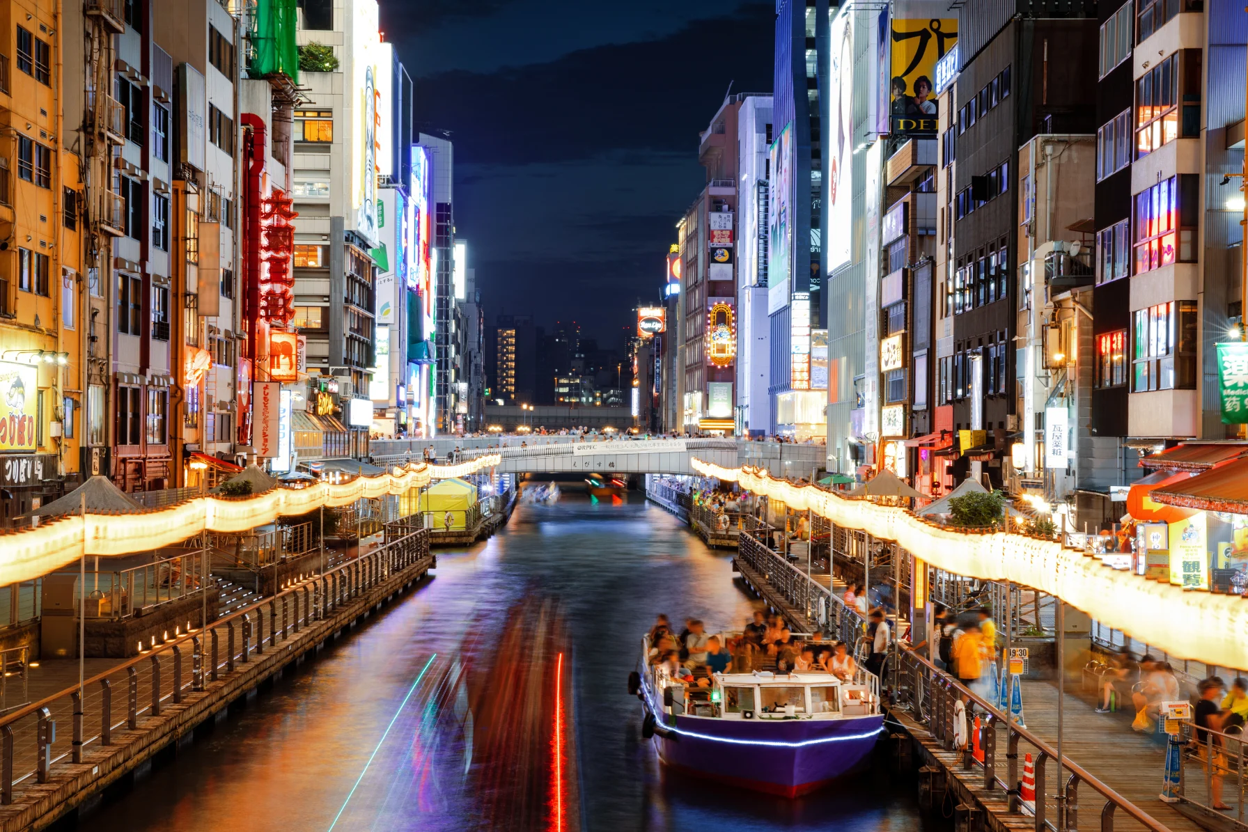 Illuminated Glico Running Man neon billboard reflecting in Dotonbori Canal with sightseeing boats in Osaka